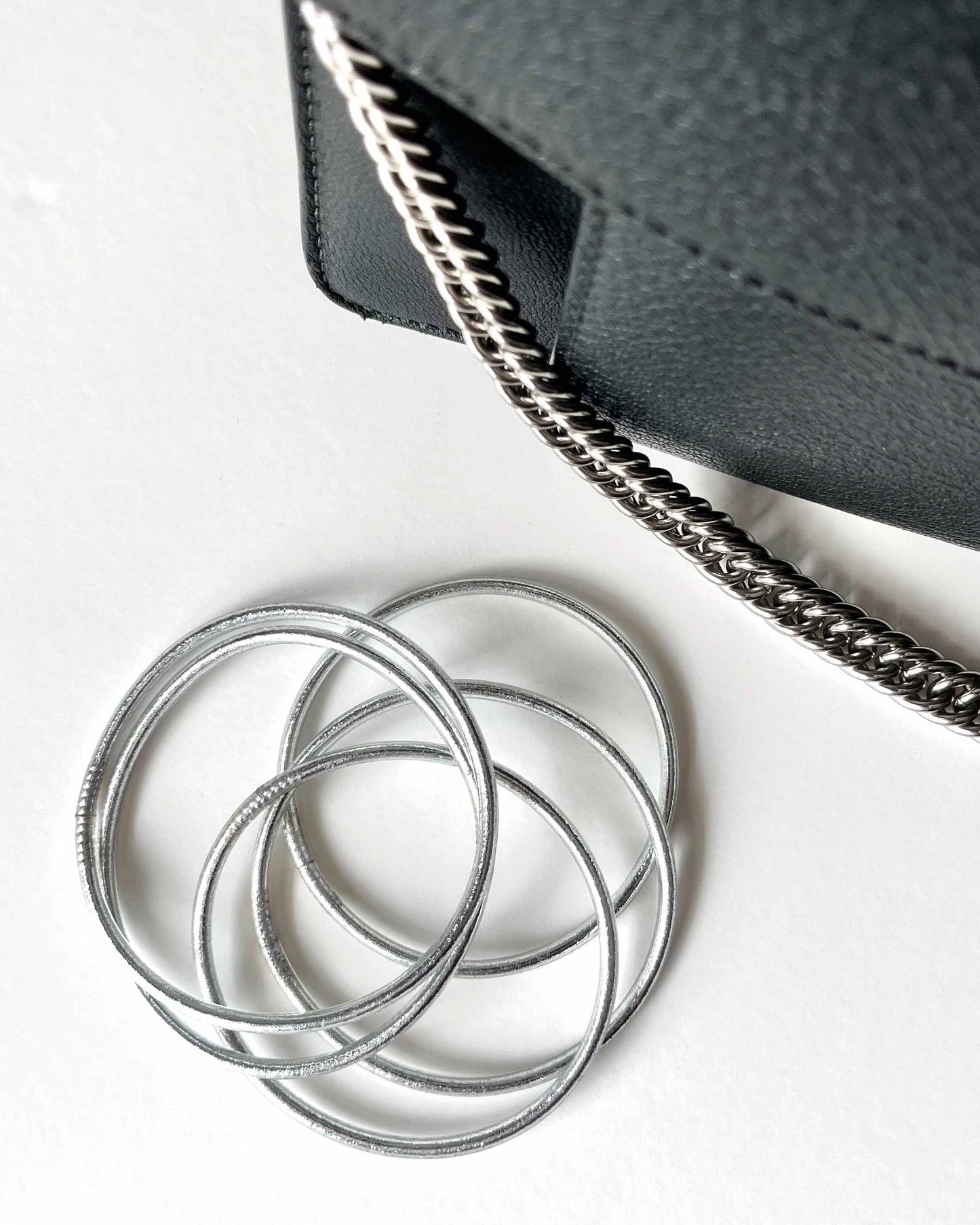 Thin Kumlai Buddhist bangles in Classic Silver, placed next to a black leather handbag.