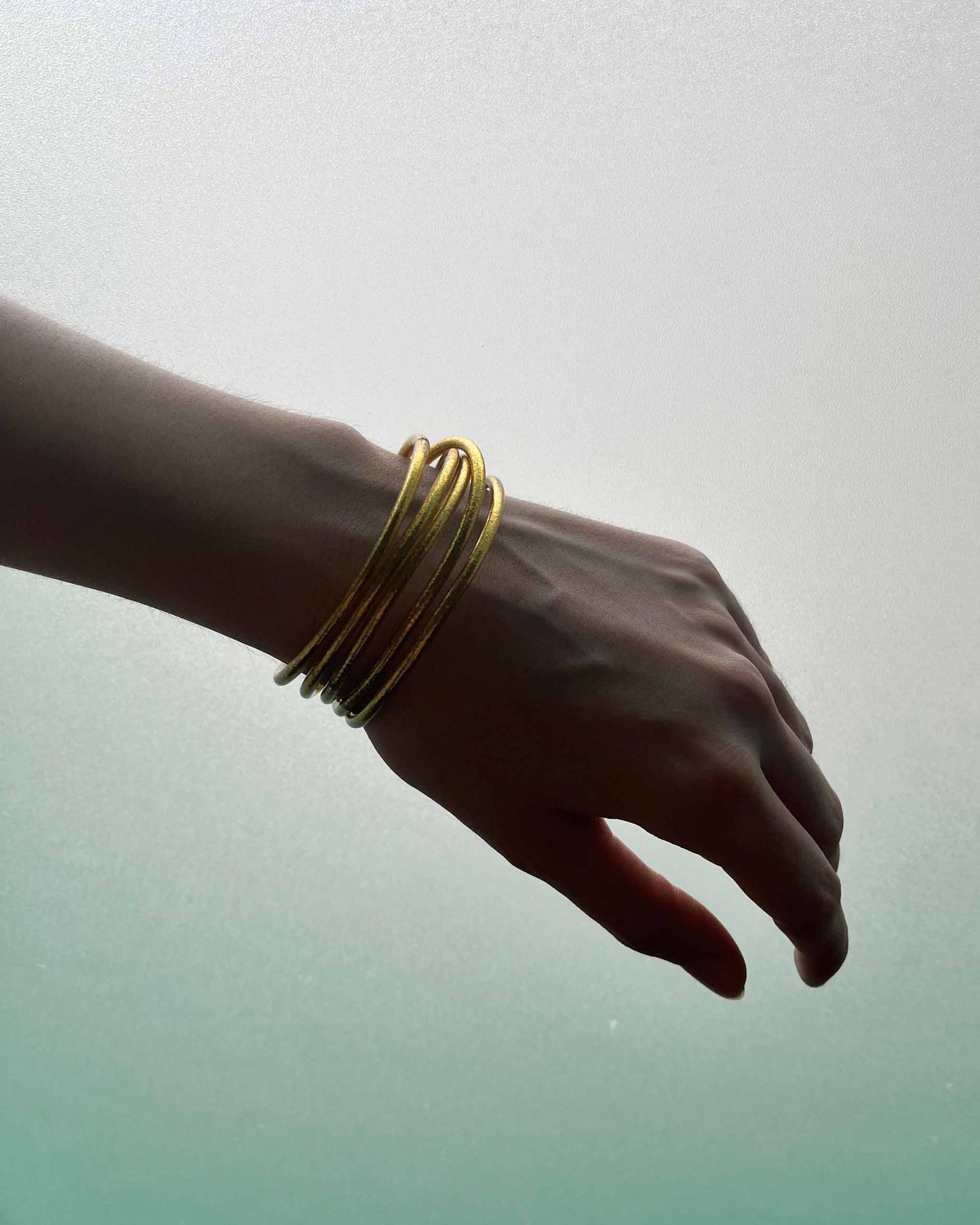 Slim Kumlai Buddhist bangles in Classic Gold, worn in front of a frosted green window.