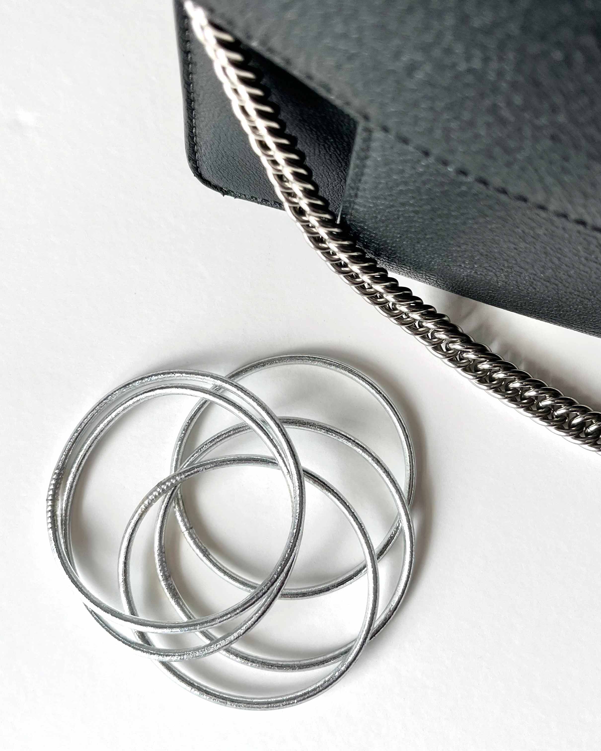 Pulseras budistas Kumlai en plata clásica, estilo fino, junto a un bolso de cuero negro.