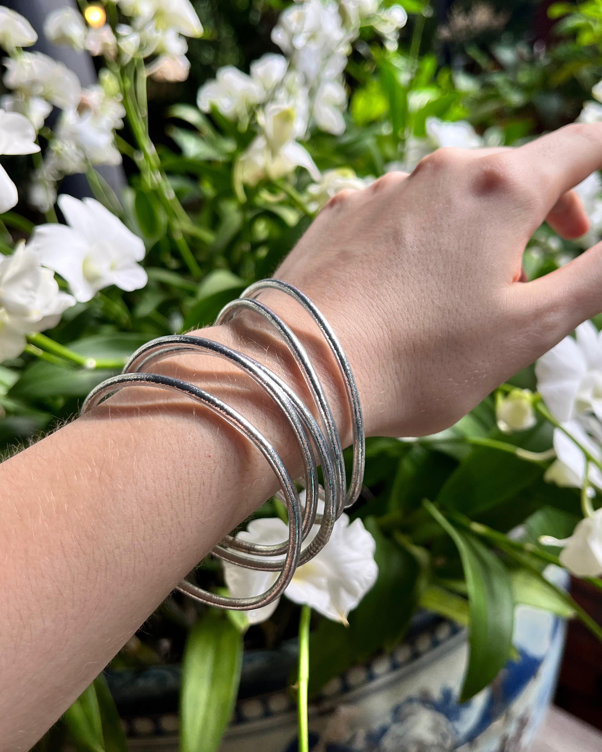 Pulseras budistas Kumlai en plata clásica, estilo fino, usadas sobre un fondo de flores blancas.