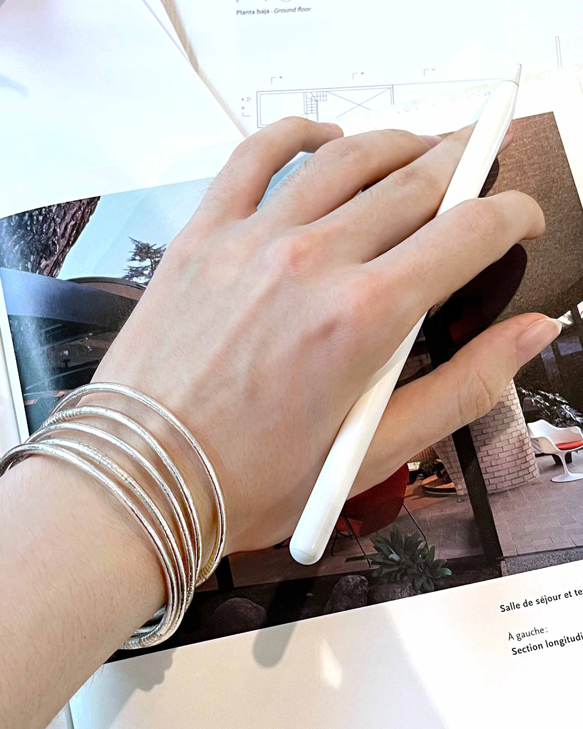 Slim Kumlai Buddhist bangles in classic silver, worn against an architecture magazine background.