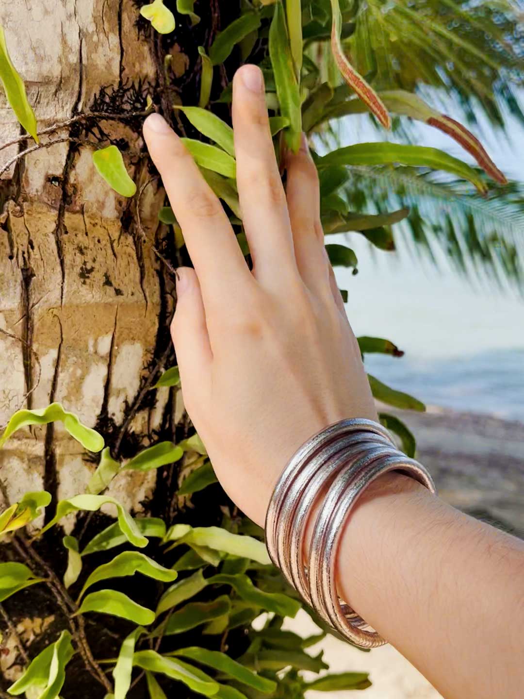Kumlai Buddhist bangles in rose silver, chunky style, against a tropical palm tree backdrop.