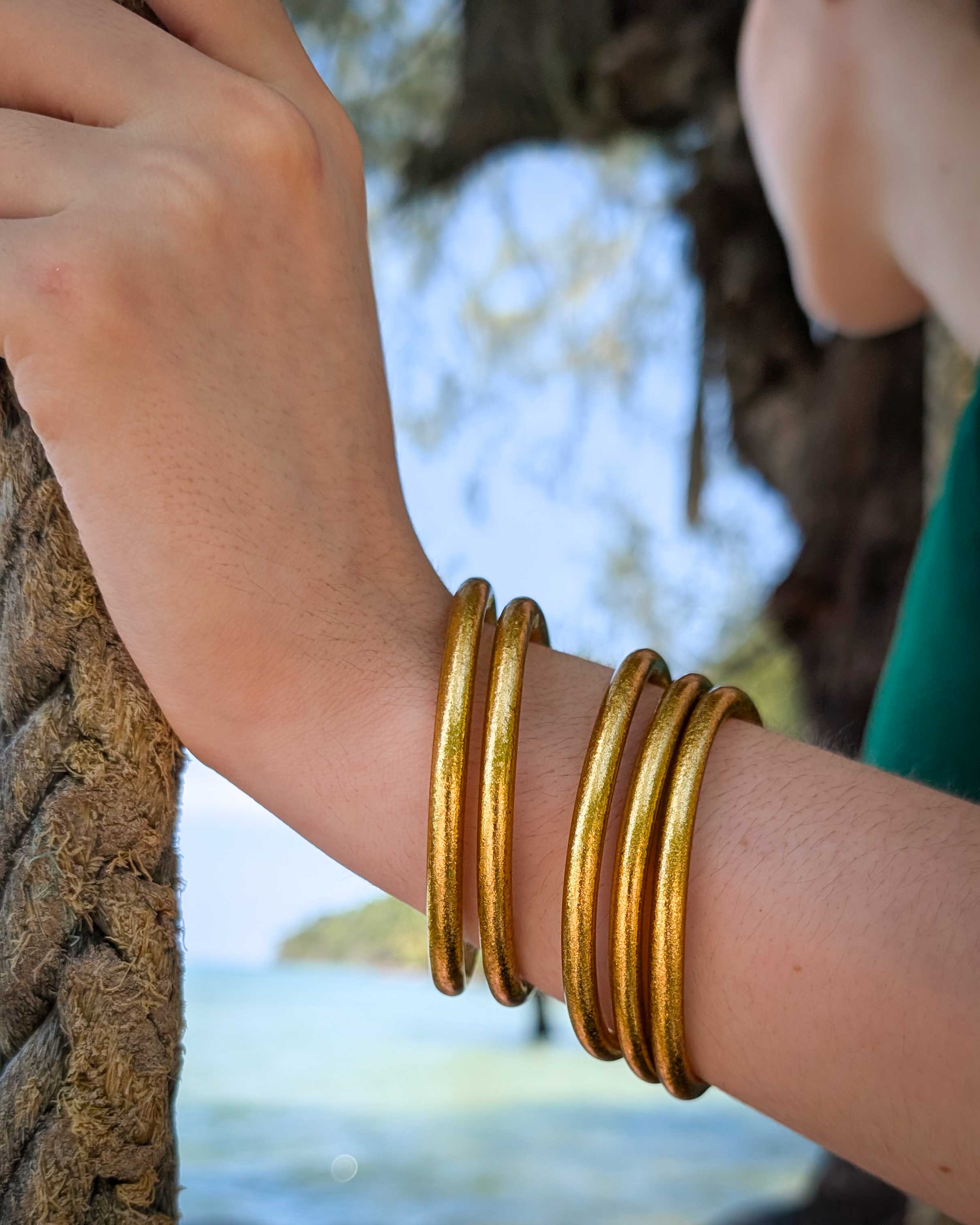 Kumlai Buddhist bangles in classic gold, chunky style, worn with a green swimming suit on a tropical sea backdrop.