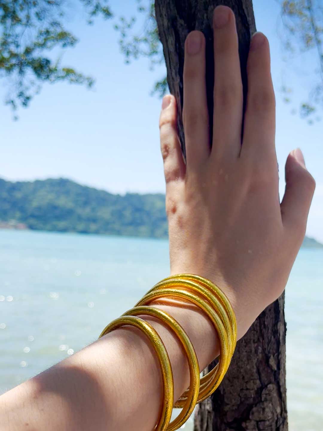 Kumlai Buddhist bangles in rich gold, chunky style, against a tropical beach backdrop.