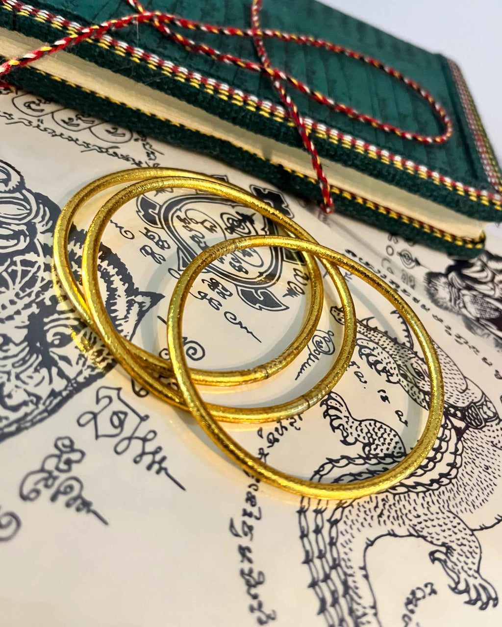 Pulseras budistas Kumlai en oro rico, estilo fino, colocadas sobre una ilustración de tatuaje tailandés.