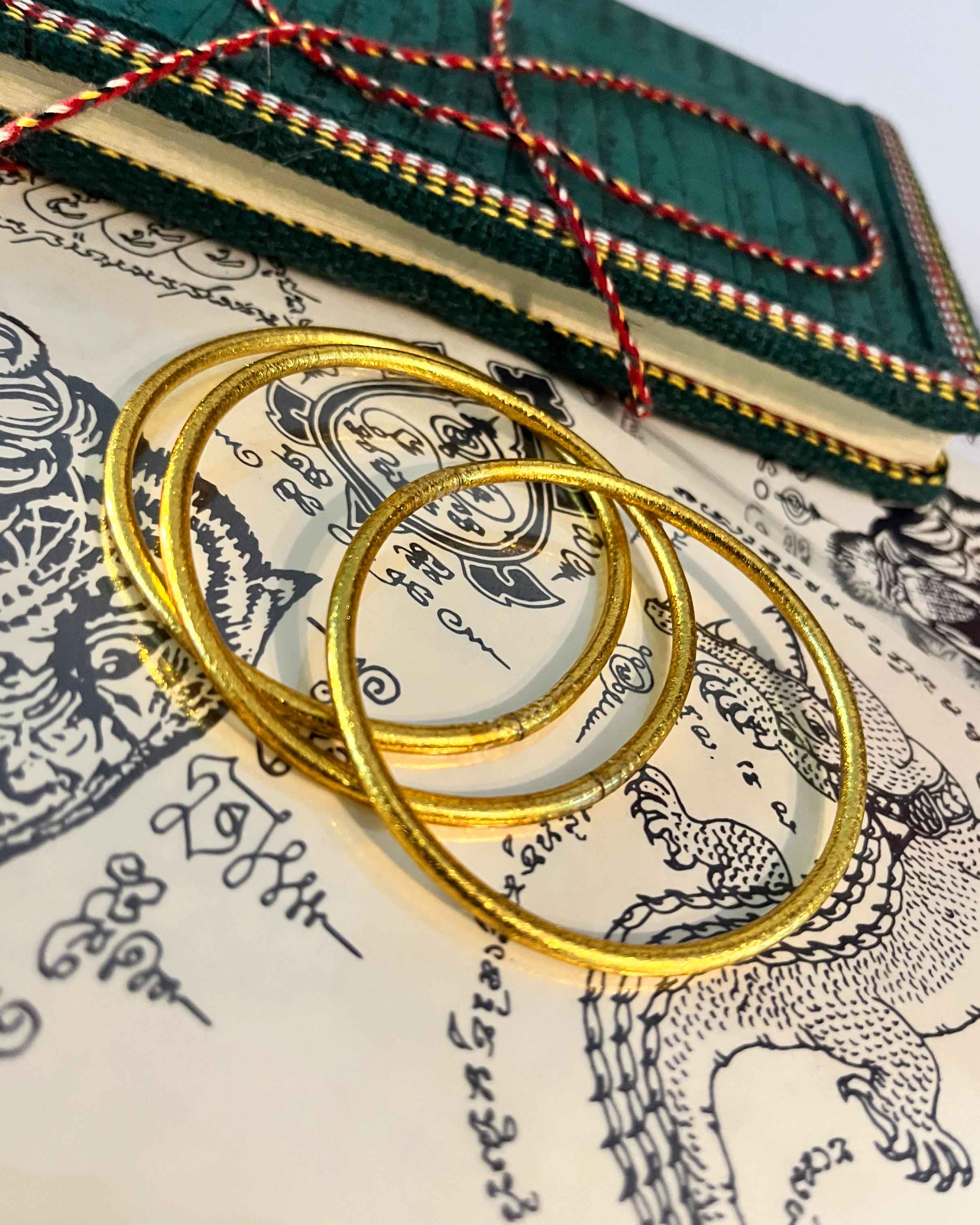Pulseras budistas Kumlai en oro rico, estilo fino, colocadas sobre una ilustración de tatuaje tailandés.
