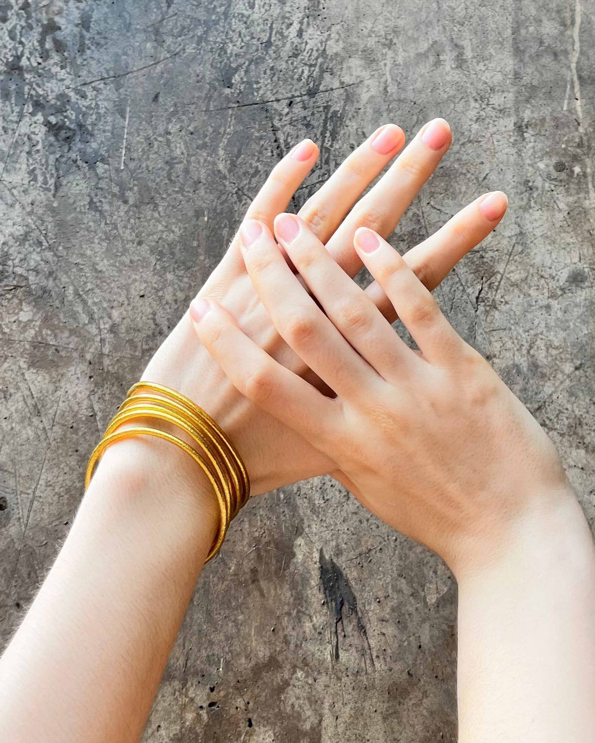 Slim Kumlai Buddhist bangles in rich gold, worn against a weathered concrete wall background.