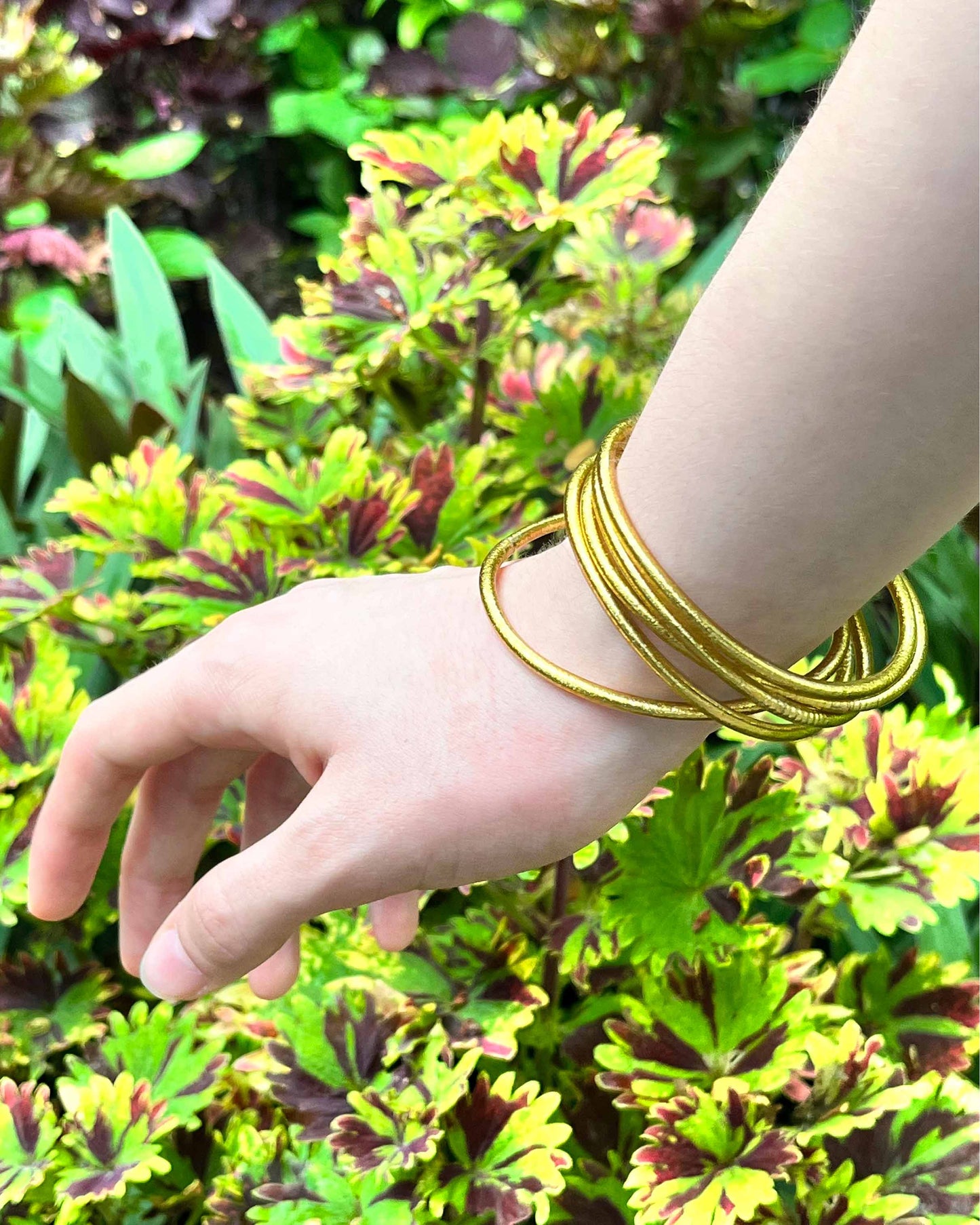 Slim Kumlai Buddhist bangles in rich gold, worn against a flower bed background.