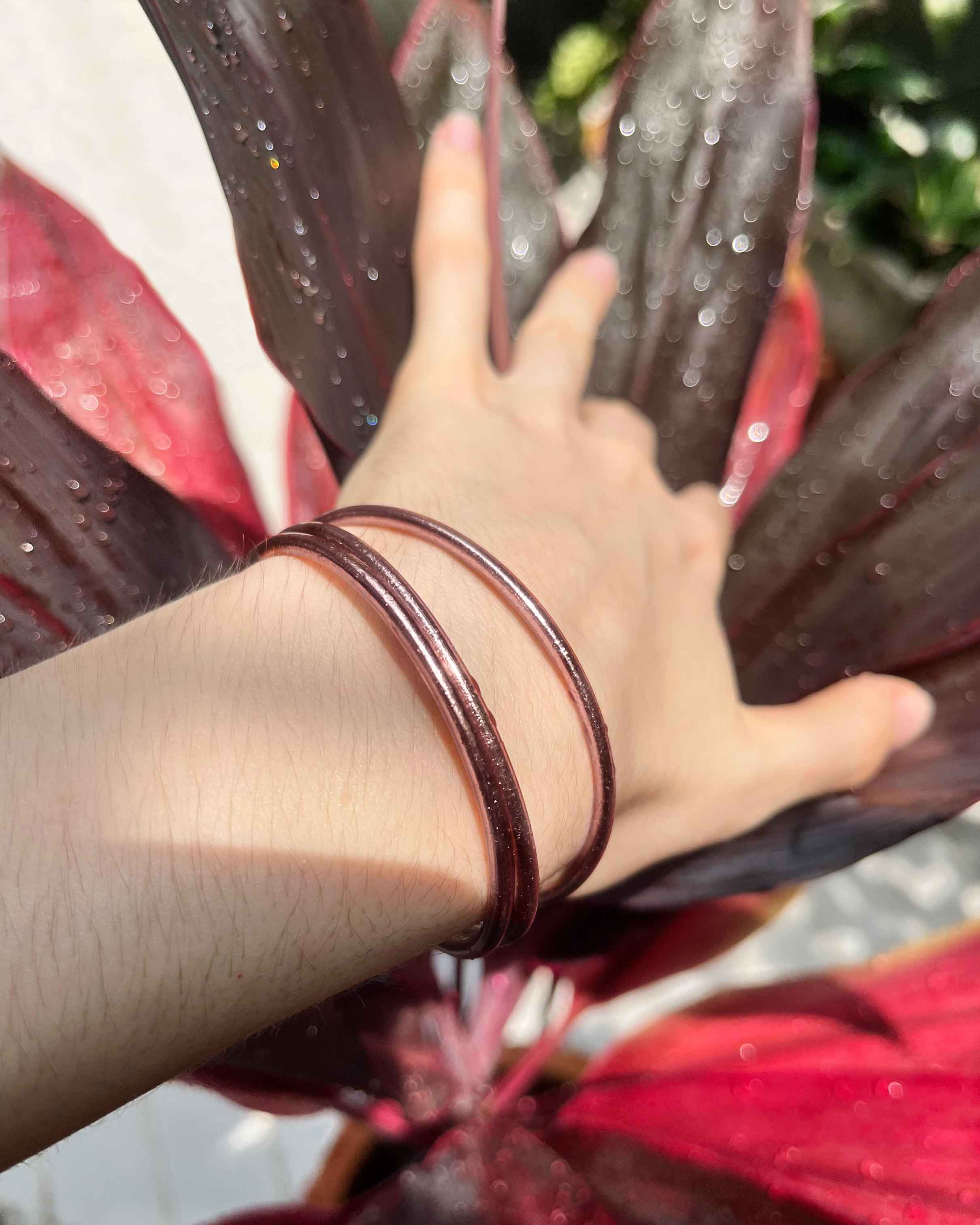 Kumlai Buddhist bangles in rose gold, slim style, worn against a red plant background.
