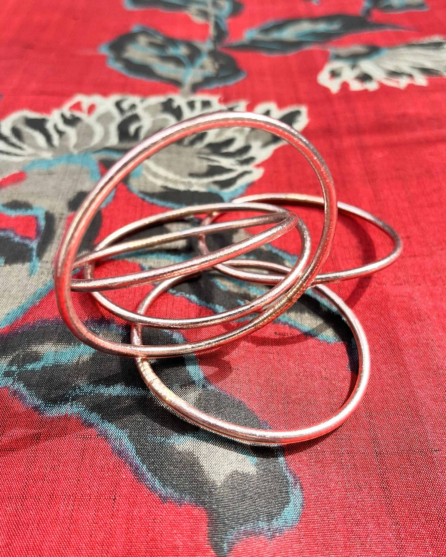 Slim Kumlai Buddhist bangles in rose gold, placed on silk fabric with floral patterns.