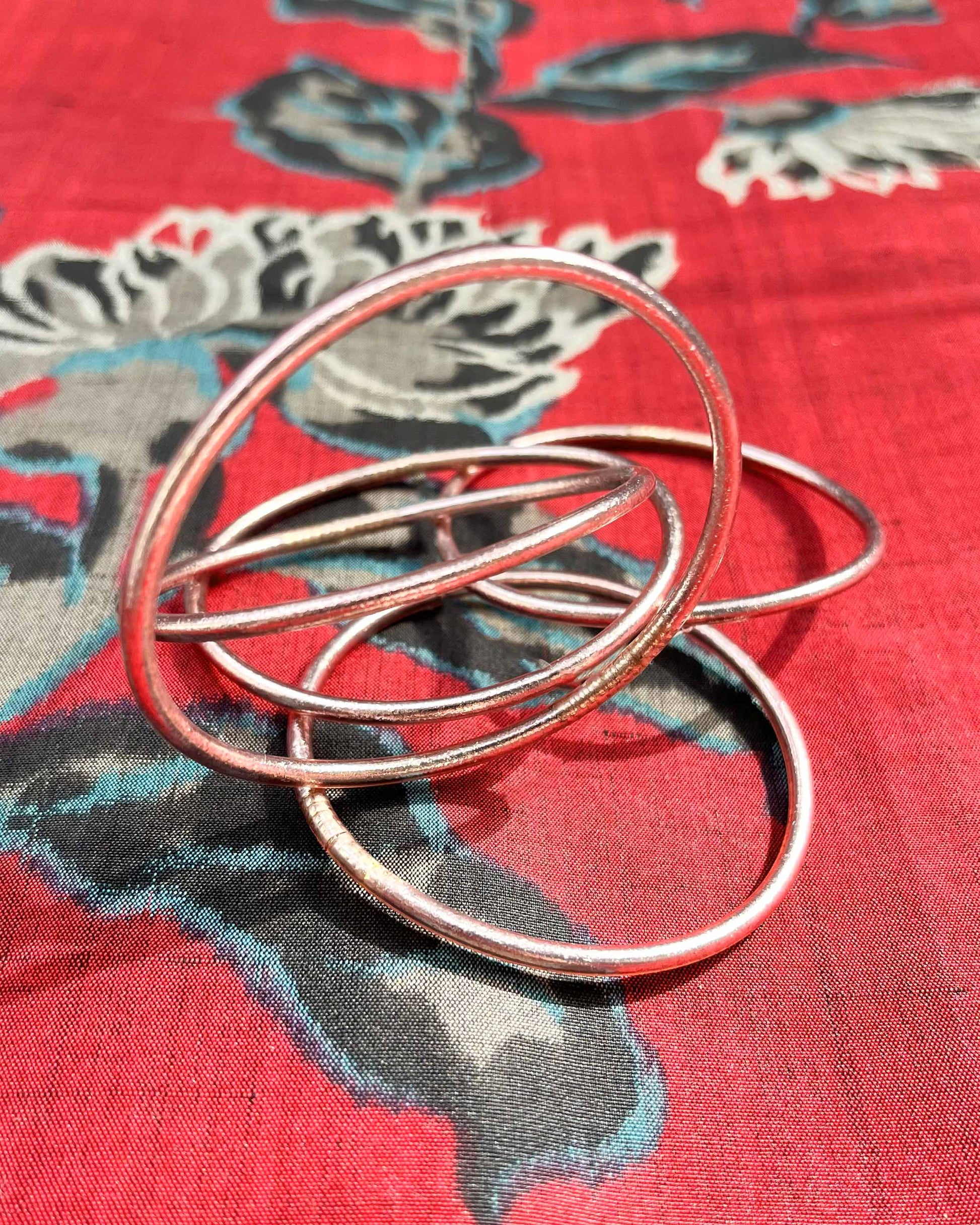 Slim Kumlai Buddhist bangles in rose gold, placed on silk fabric with floral patterns.